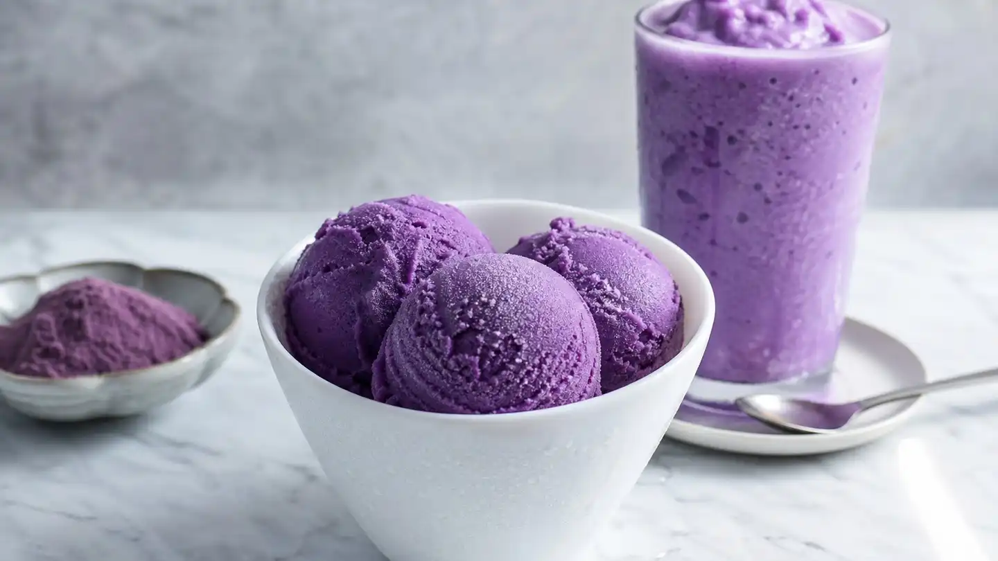 Three scoops of purple yam ice cream in a white bowl beside a tall glass of purple frozen shake on a marble counter
